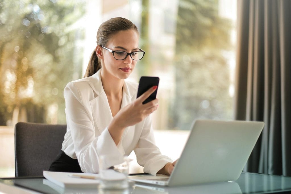 Professional woman using smartphone while working on laptop in modern office.